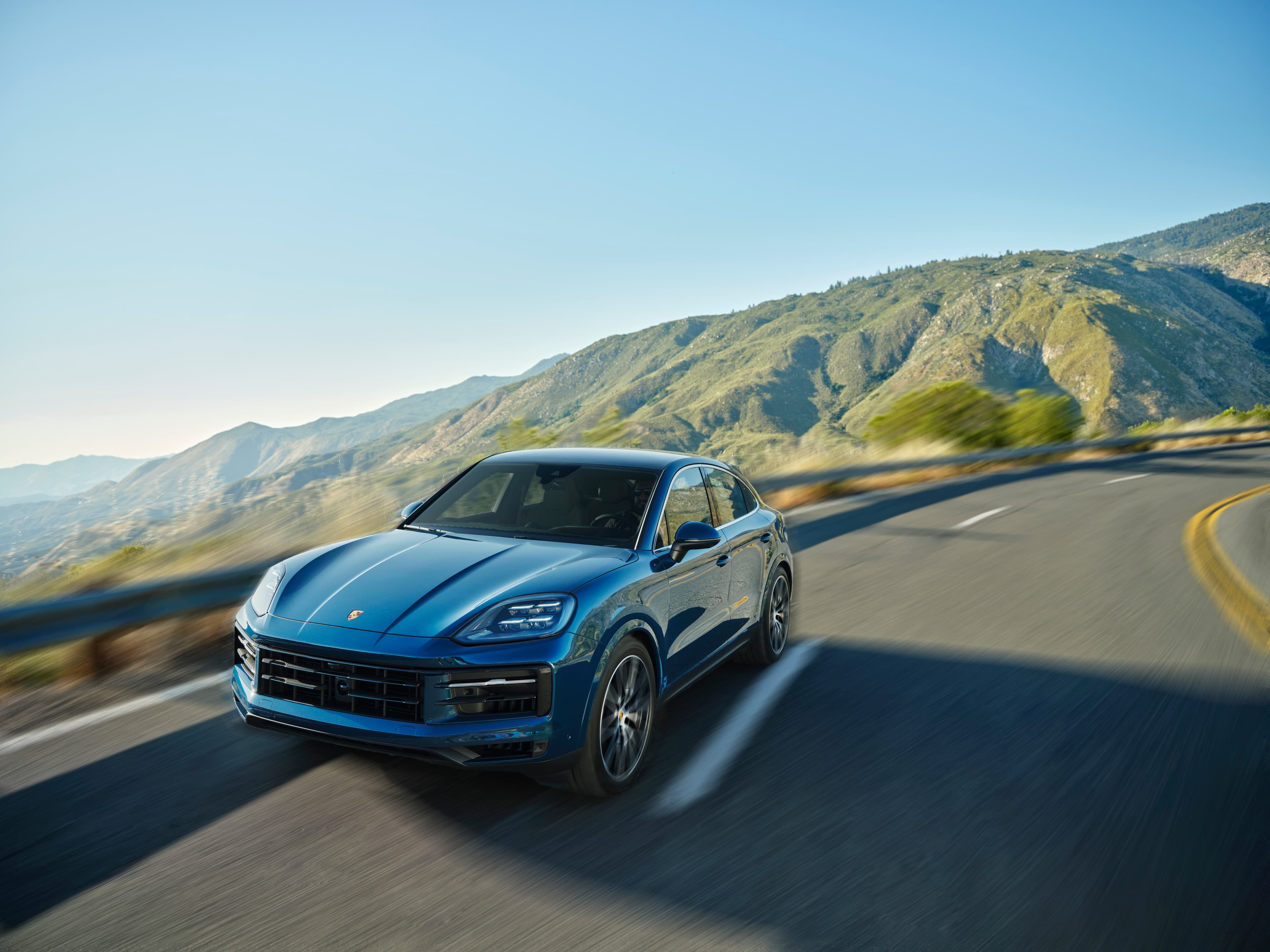 A blue 2023 Cayenne S Coupé drives on a scenic mountain road, with blurred motion and green hills in the background under a clear blue sky.